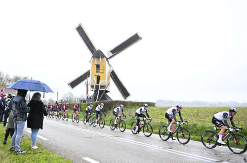 Mensen onder een paraplu kijken hoe wielrenners tijdens de Amstel Gold Race van 2023 door een regenachtig Limburgs landschap koersen. 